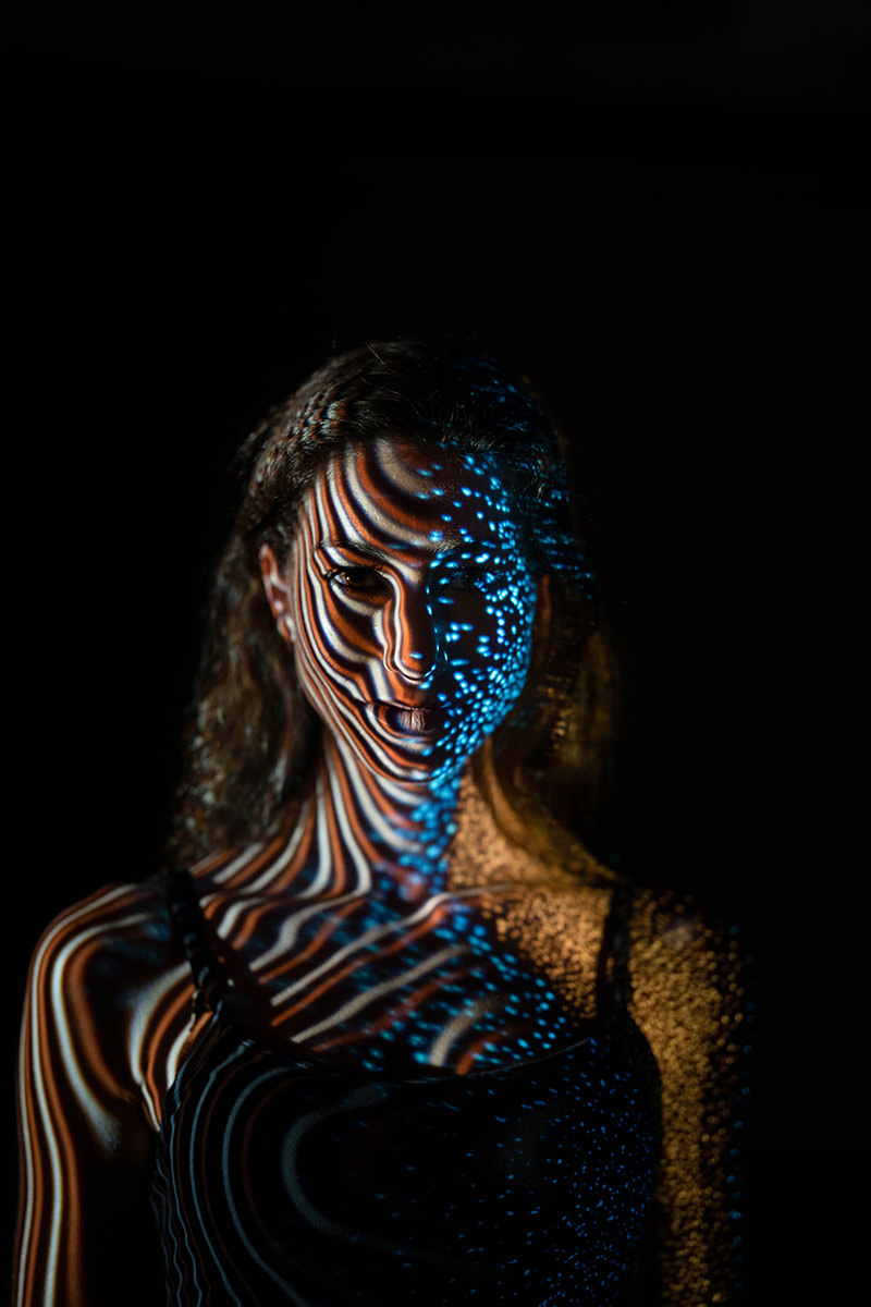 Woman with projected abstract patterns of blue dots and orange and black striped lines on her face and shoulders, set against a black background