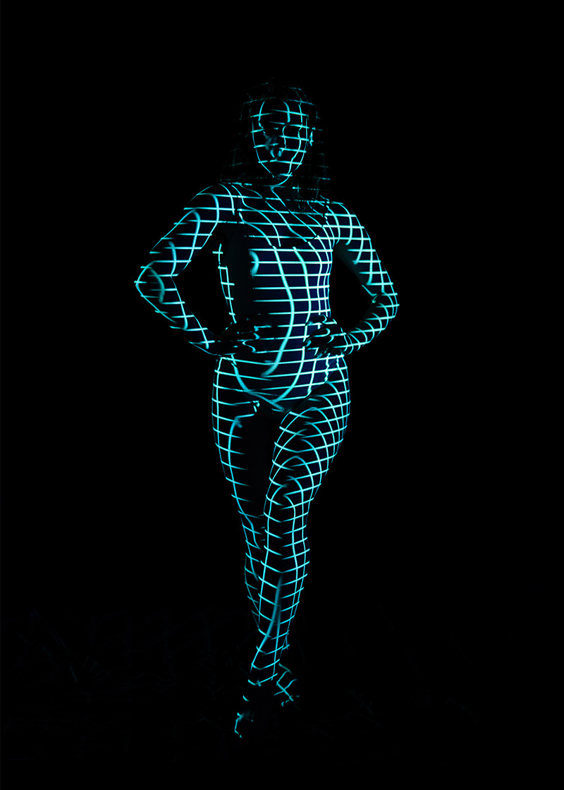 Person standing with hands on hips, covered in a glowing blue grid pattern projected on their body and face against a black background
