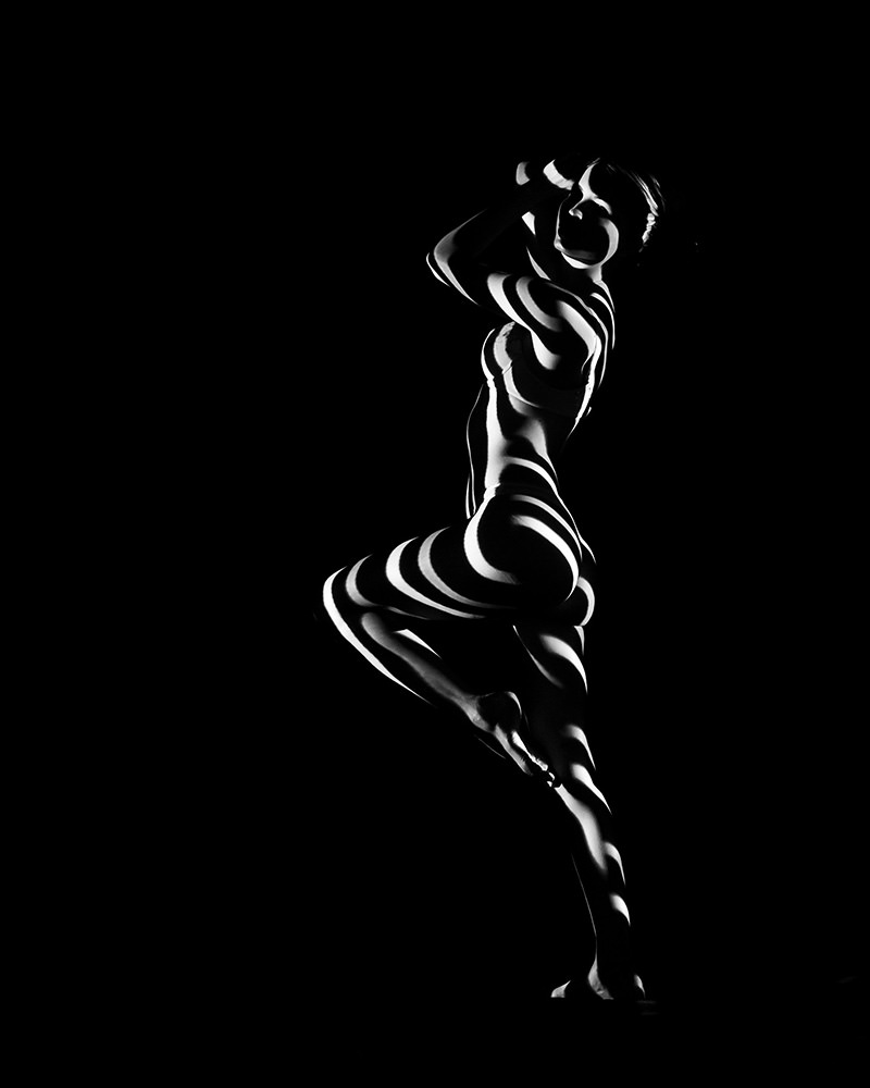 Nude person posed with one leg raised, illuminated by dramatic black and white striped light patterns against a dark background