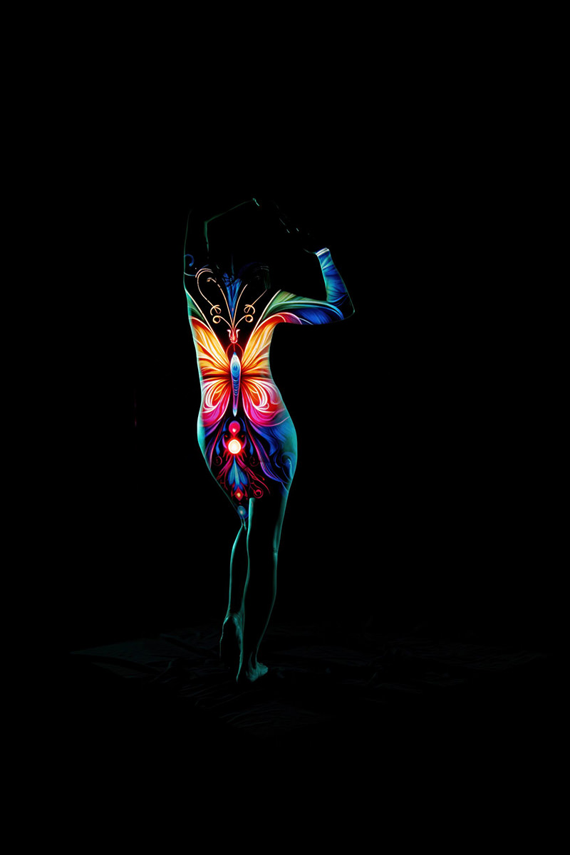 Silhouette of a person with colorful butterfly design painted on their back, arms, and legs, standing on tiptoe with arms raised, against a black background