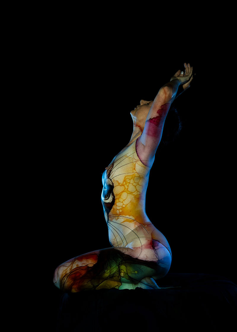 Person kneeling with head tilted back and arms raised, body covered in colorful abstract art and patterns, set against a black background