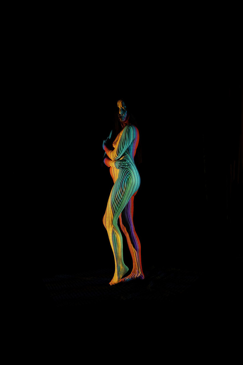 A standing nude model with a colorful checkered and striped pattern projected onto their body, set against a black background. The patterns create a multi-colored visual effect with stripes of green, orange, purple, and blue wrapping around the figure.