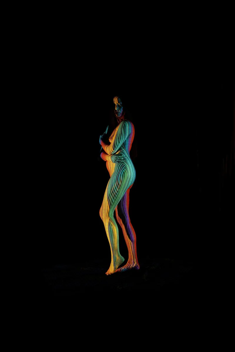 A standing nude model with a colorful checkered and striped pattern projected onto their body, set against a black background. The patterns create a multi-colored visual effect with stripes of green, orange, purple, and blue wrapping around the figure.