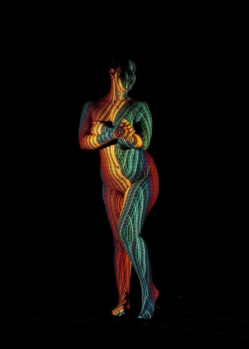 Naked person standing against a black background with a colorful pattern of small dots projected on their body, creating an artistic display of orange, red, yellow, blue, and green hues.
