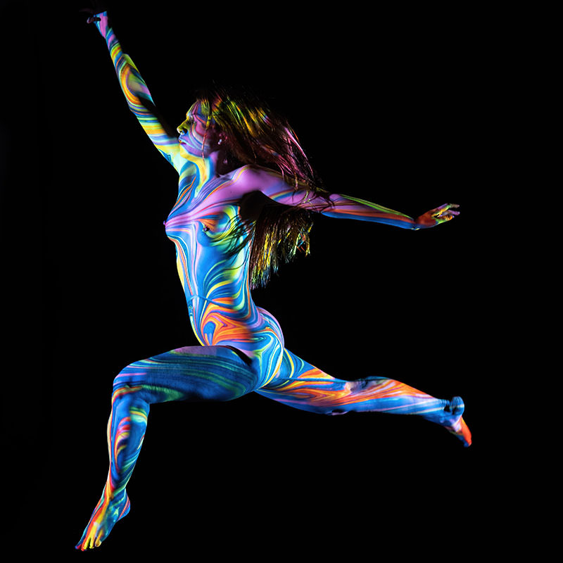Nude person mid-air in a dynamic leap, covered in vibrant swirling neon patterns of blue, orange, yellow, and green against a black background