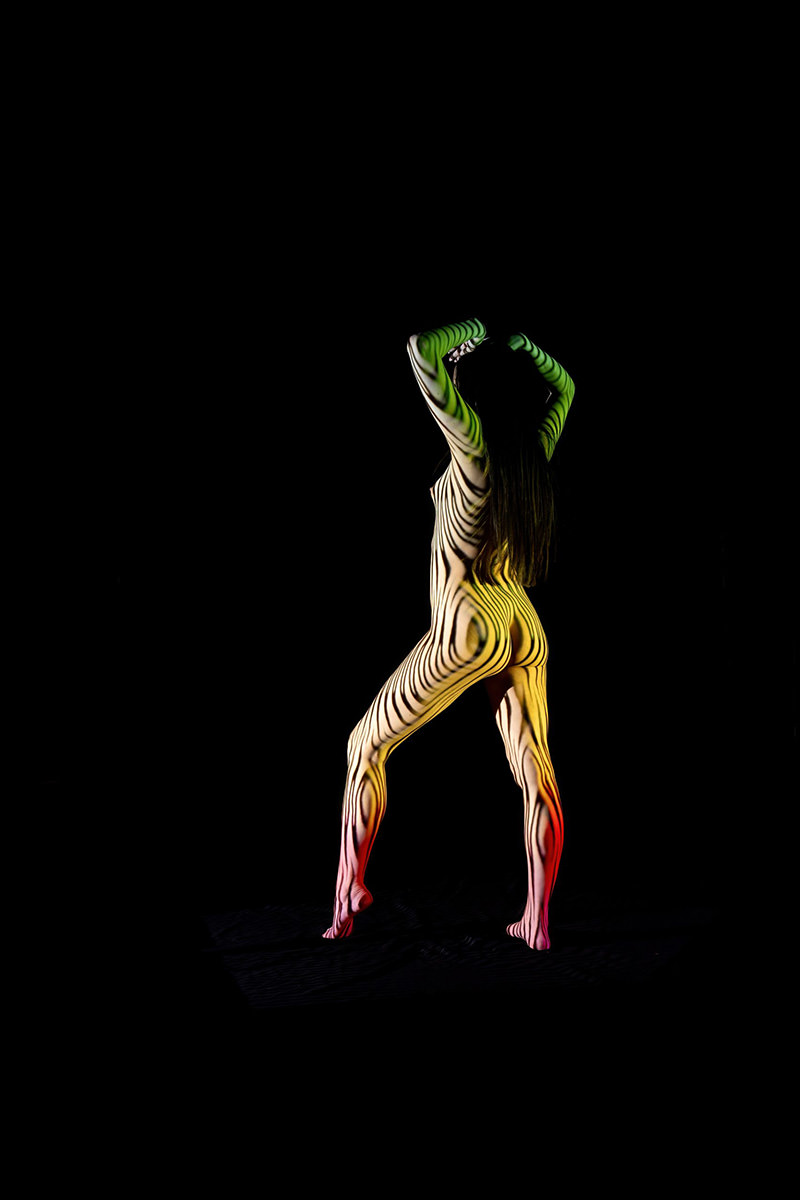 Nude person standing with arms raised, body covered in swirling black stripes and colorful light projections against a black background