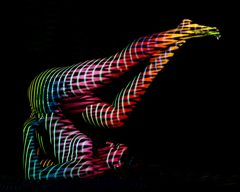 Human body contorted with legs extended upwards, covered in vibrant striped light patterns against a black background