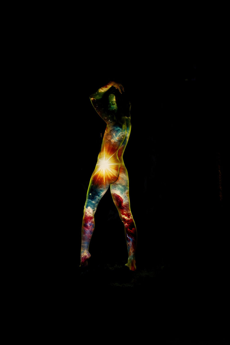 Person standing with hands raised, body painted or projected with colorful cosmic imagery including stars, nebulae, and a bright glowing star at the lower back against a black background