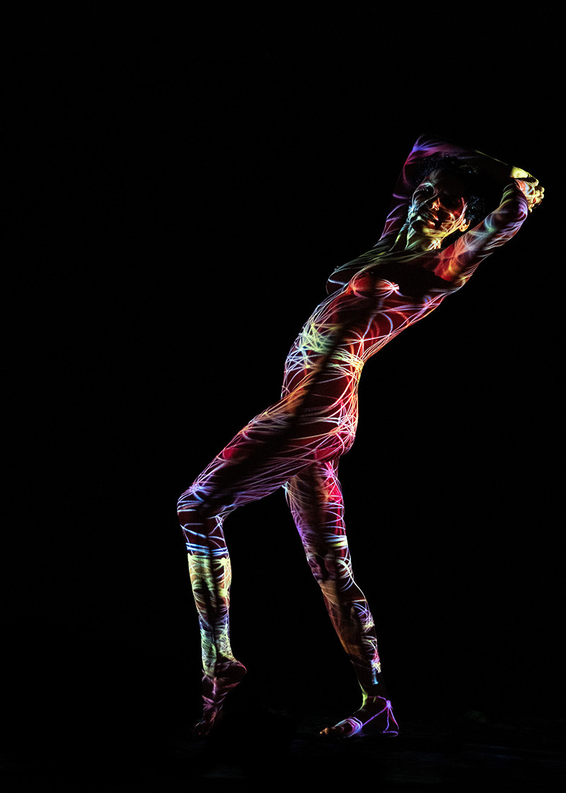 A person standing on tiptoe against a black background, their body covered in colorful, swirling light projections creating dynamic patterns.