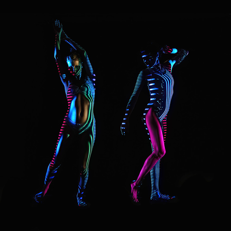 Two dancers posing against a dark background with blue, pink, and green striped light patterns projected onto their bodies, creating a vibrant and artistic effect.
