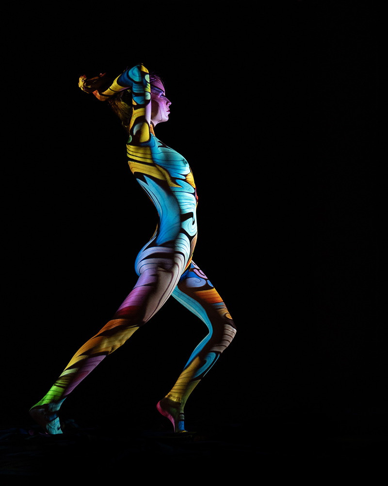 Person standing in a dynamic pose with arms raised, their entire body covered in intricate, colorful, swirling patterns against a black background