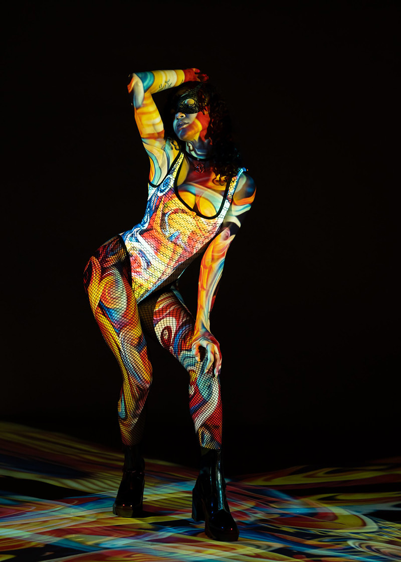 Person in colorful patterns projected onto body wearing a fishnet bodysuit, high platform boots, and a black mask, posing against a dark background with swirling bright colors projected on the floor.