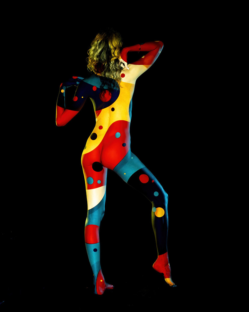 Back view of a woman with long curly hair posing with one leg lifted, covered head to toe in vibrant body paint with abstract patterns of red, yellow, blue, black, and white on a black background
