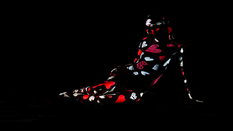 A person sitting in the dark with red, white, and black heart patterns projected across their body and clothes.