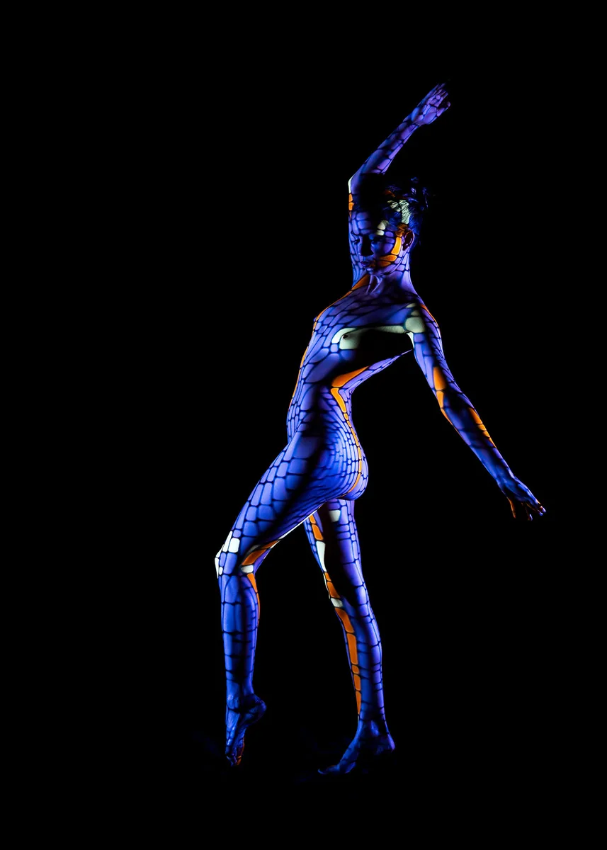 A nude person posing gracefully with arms raised, covered in a mosaic-like pattern of blue, white, and orange lights against a black background