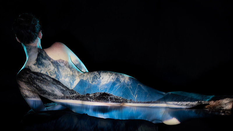 Reclining nude person with a detailed mountain landscape projection cast on their back and legs against a black background