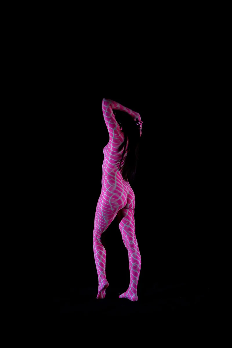 Nude person standing with one leg slightly raised and arms raised behind head, covered in glowing pink geometric grid patterns against a black background