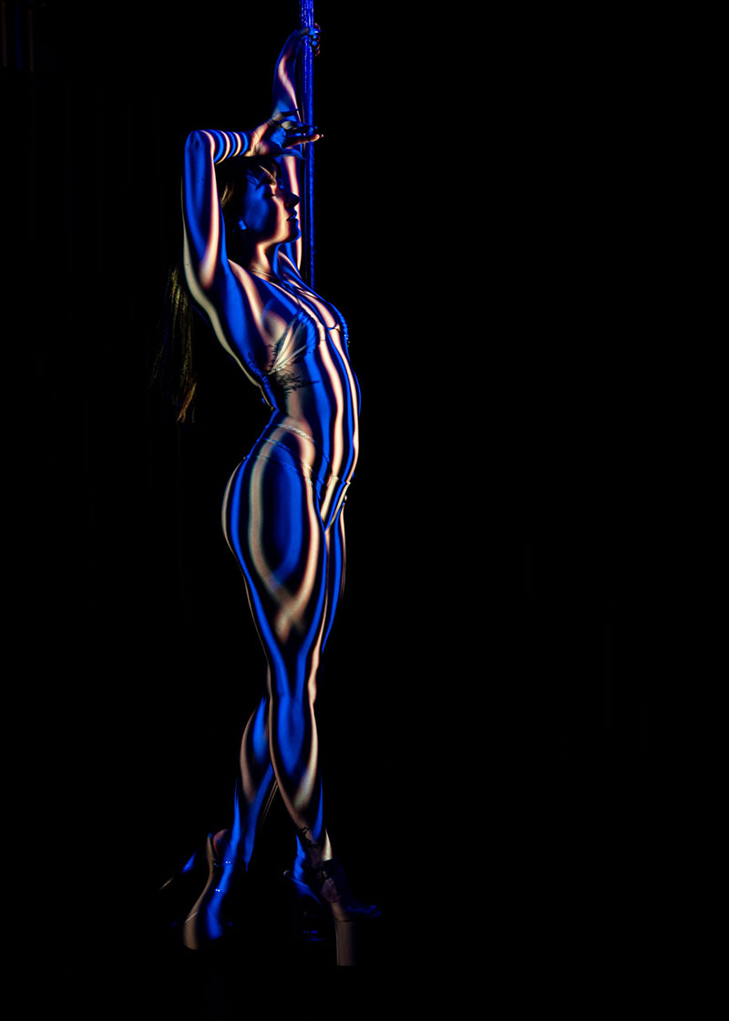 A woman posed against a pole with arms raised, illuminated by blue and white vertical striped lighting that contours her body, set against a dark background.