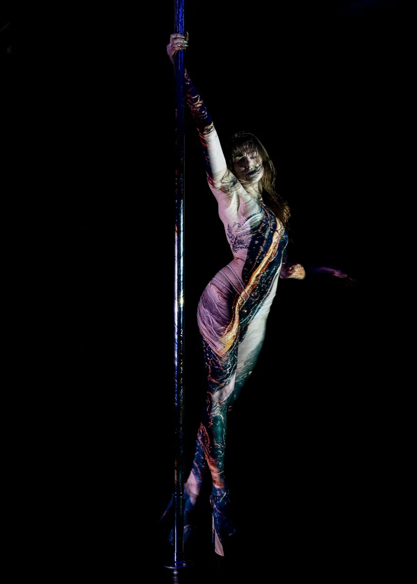 Person performing a pole dance illuminated by colorful abstract projections on their body against a dark background