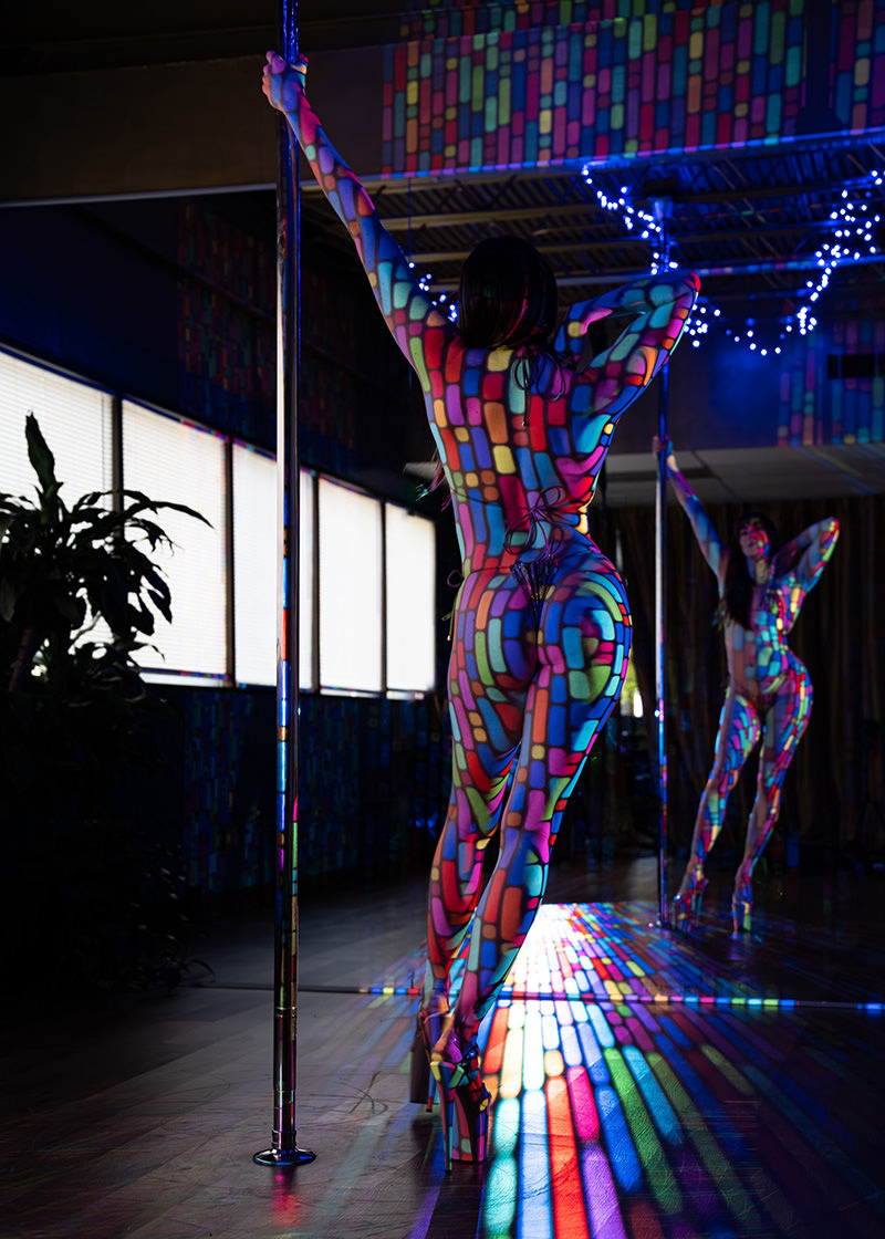 A woman poses next to a vertical pole in high heels with multicolored light patterns projected on her body and the surrounding floor. A mirror reflects her full figure posing with one hand on her head and the other on the pole.