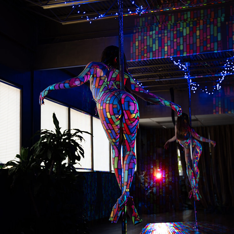 Pole dancer wearing high heels with colorful, geometric light patterns projected on their body, standing in a dance studio with mirrored walls and large windows, surrounded by plants and blue fairy lights.