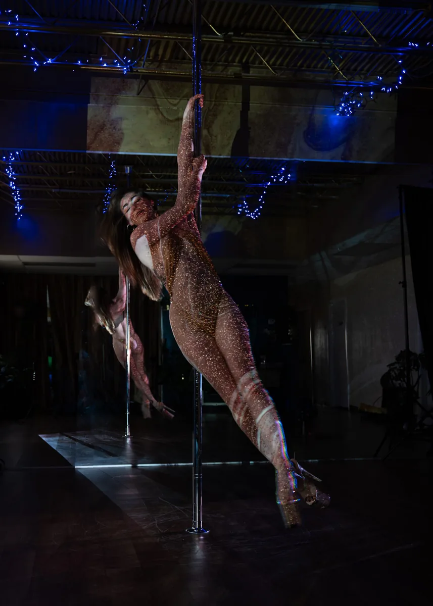 A person performing a pole dance move with one hand gripping the pole, body extended horizontally, wearing a glittery bodysuit. Blue string lights hang from the ceiling and a large mirror reflects the pose.