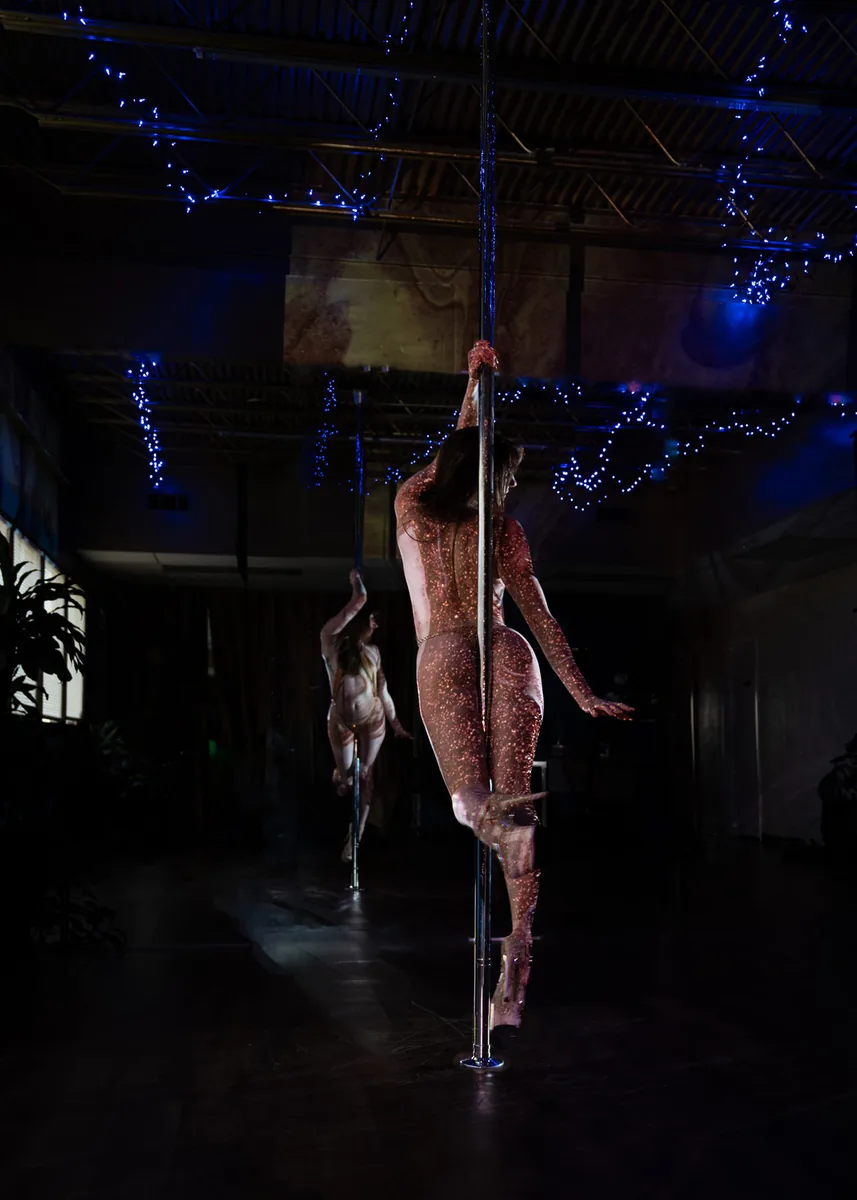 A woman performing pole dance moves in a dark studio with blue string lights on the ceiling, her body covered in small glowing dots, and a large mirror reflecting her image.