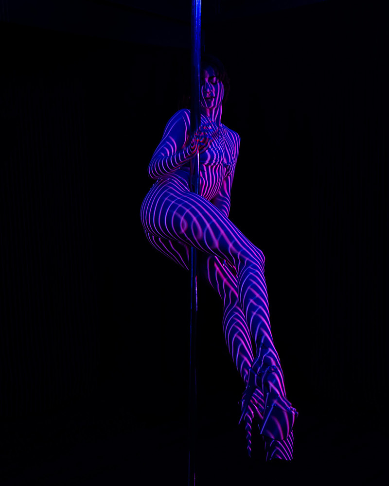 A person performing pole dance against a black background, illuminated by purple and blue striped light patterns creating an artistic visual effect on their body and face. They are wearing high heels and posing gracefully on the pole.