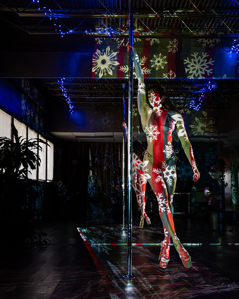 Woman in bikini and high platform heels dancing on a pole with red and white snowflake patterns projected across her body and the surrounding room, including blue string lights hanging from the ceiling.