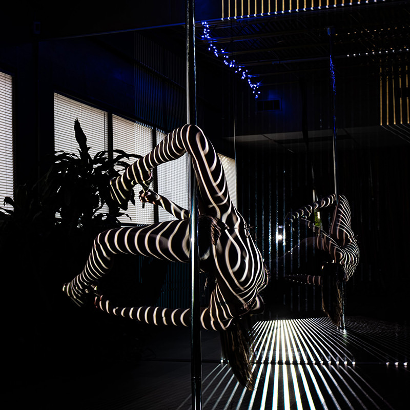 Person performing a pole dance move with bold horizontal light stripes projected over their body, bending backward gripping the pole, mirrored in the background.