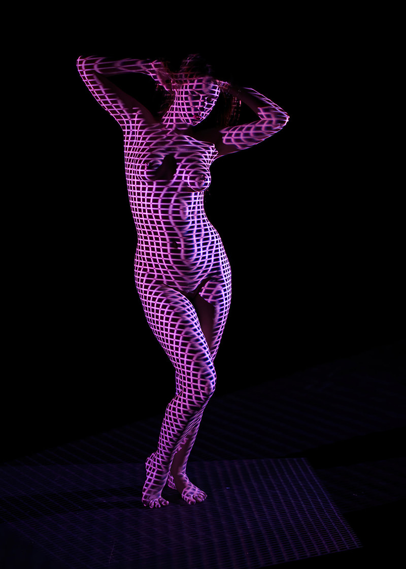 Nude person posed with arms raised, covered in a purple grid pattern projection, standing against a black background