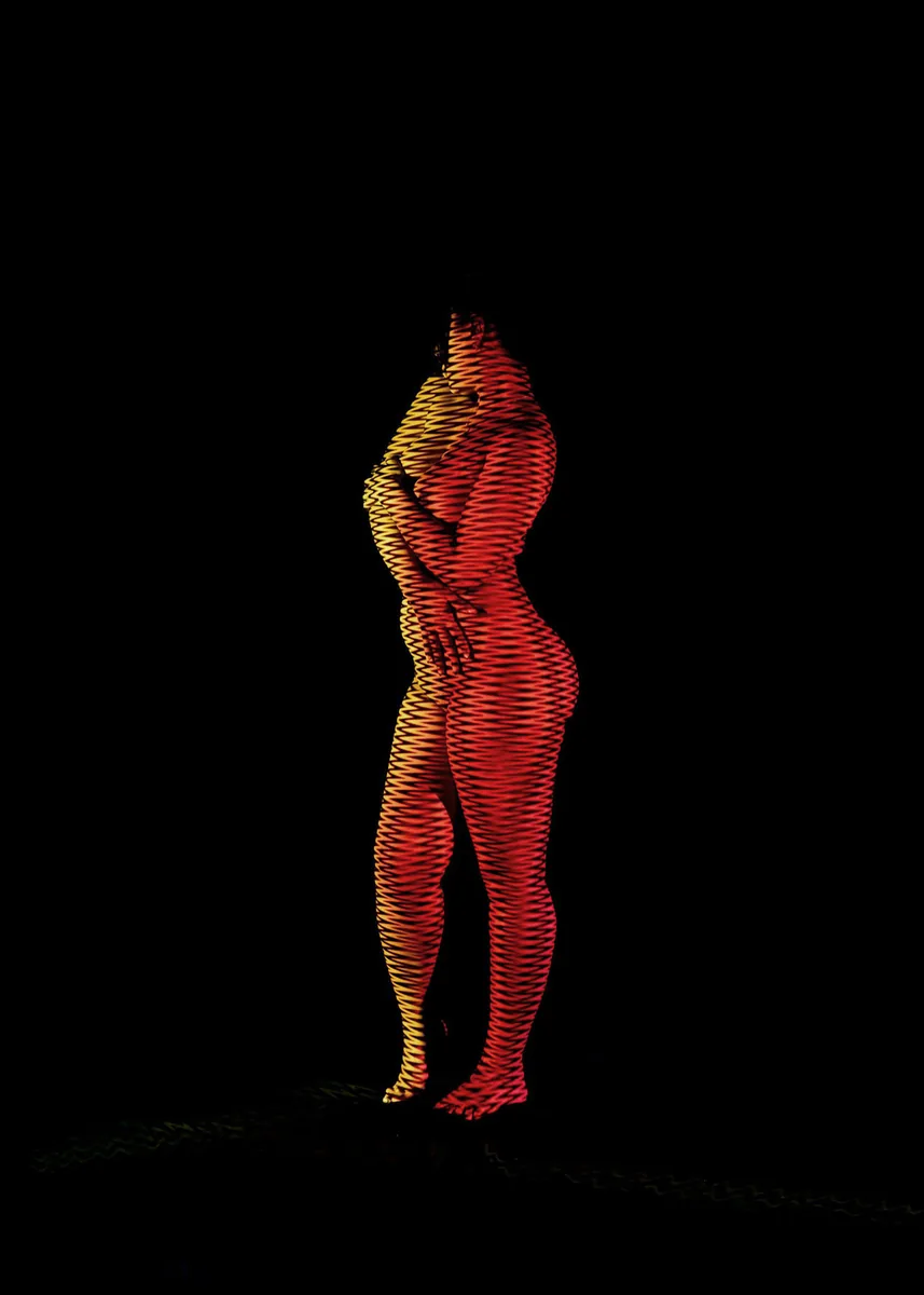 Nude standing person with arms crossed, illuminated by red and yellow zigzag light projections against a black background