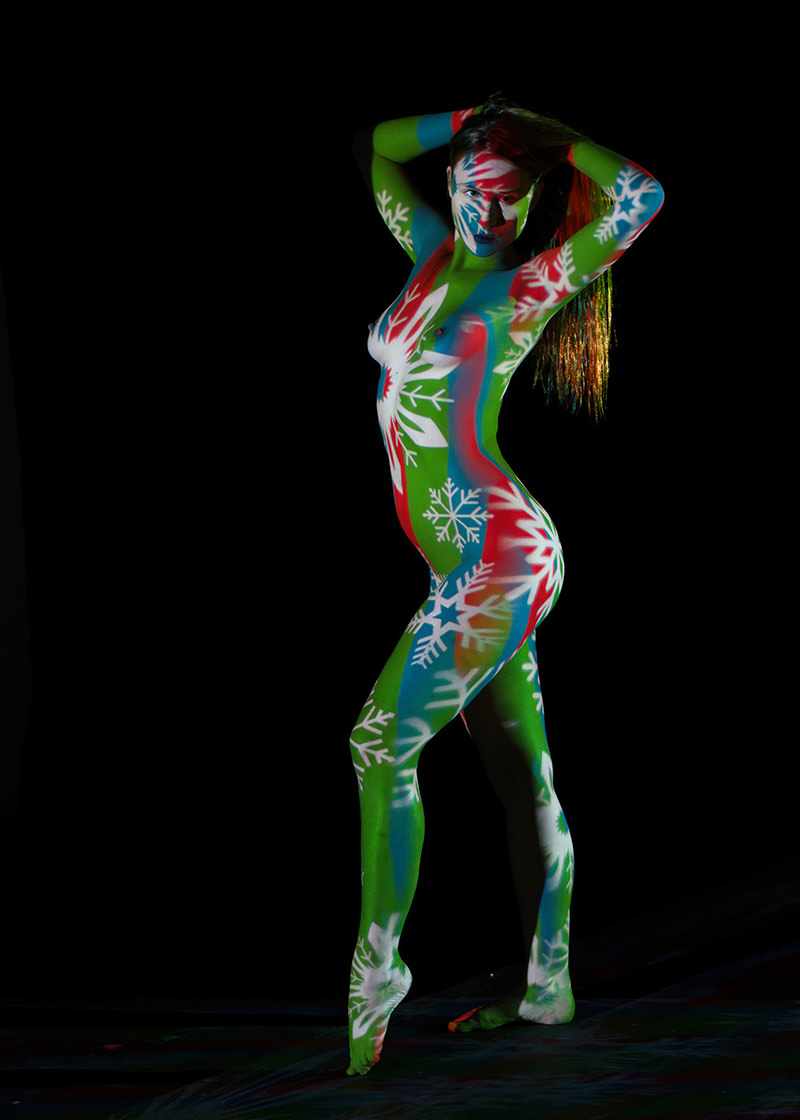 A person standing on toes with arms lifted behind head, body painted in vibrant green, red, blue, and white colors featuring large snowflake patterns against a black background.