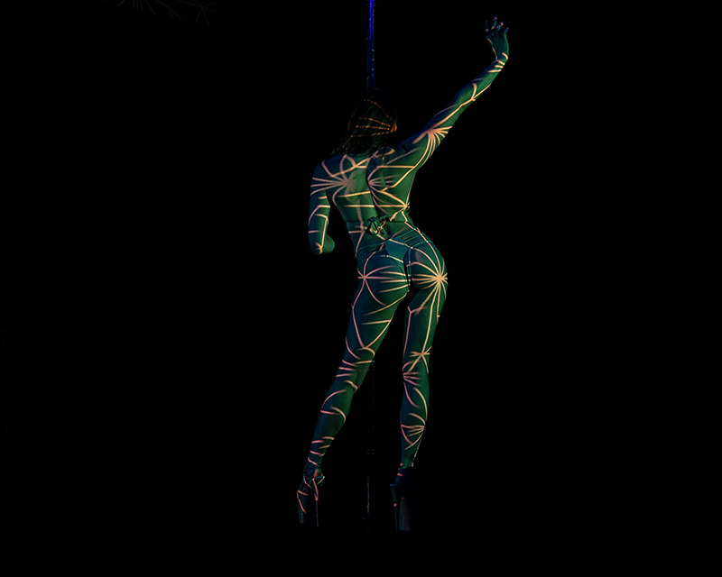 Back view of a person standing on platform heels with raised arm, covered in light projections creating a pattern of curved lines and star shapes on their body against a black background