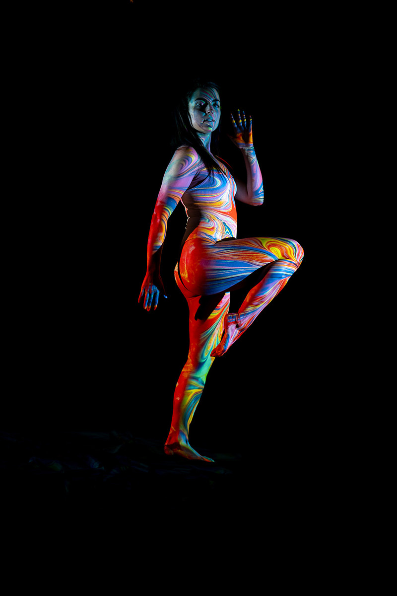 Woman balancing on one leg with the other leg lifted, covered in vibrant swirling colorful patterns against a black background