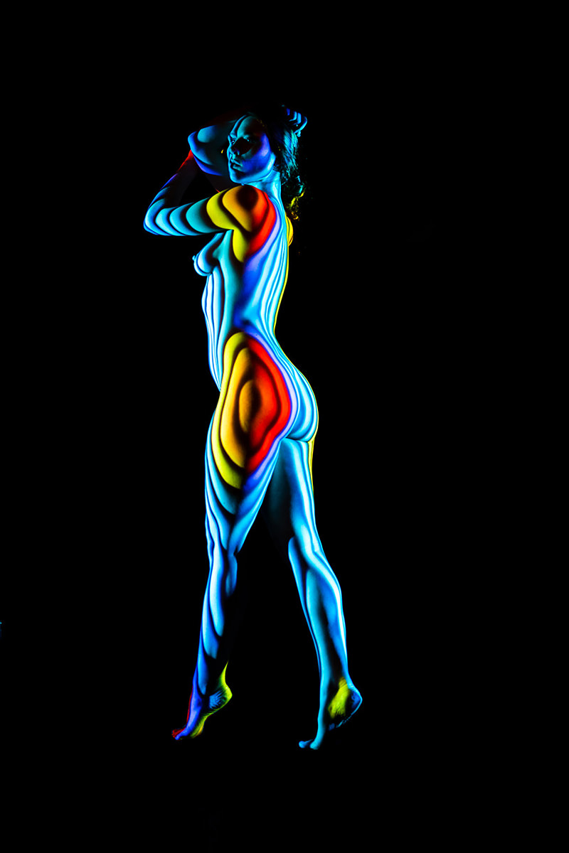 Person standing with legs slightly crossed, covered in colorful light patterns featuring blue, yellow, red, and white stripes against a black background