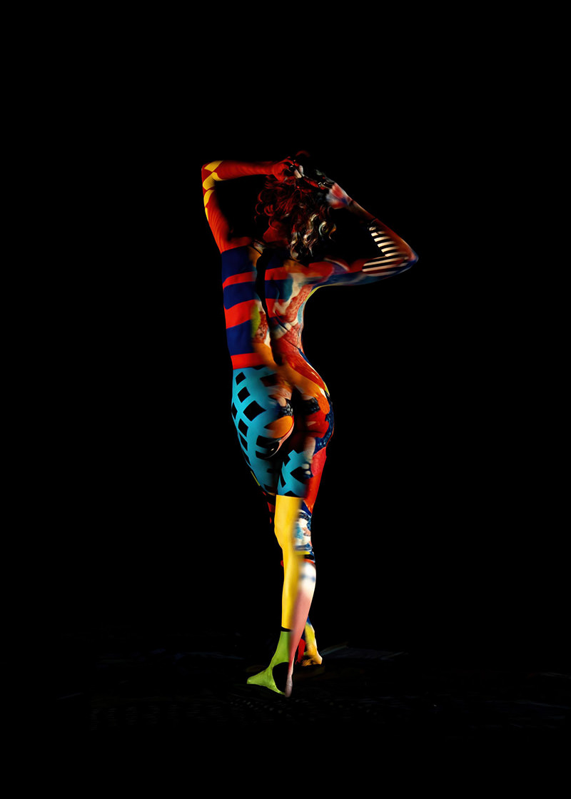 Person standing with back facing, covered in colorful abstract body paint with red, blue, yellow, and black patterns on a black background