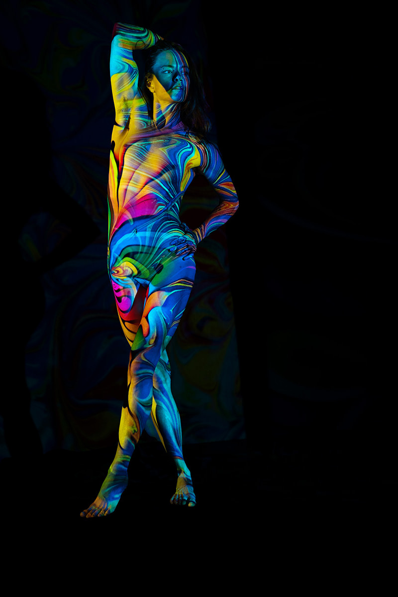 Person standing with one arm raised and the other on hip, covered in swirling projected blue, yellow, red, and green patterns against a black background