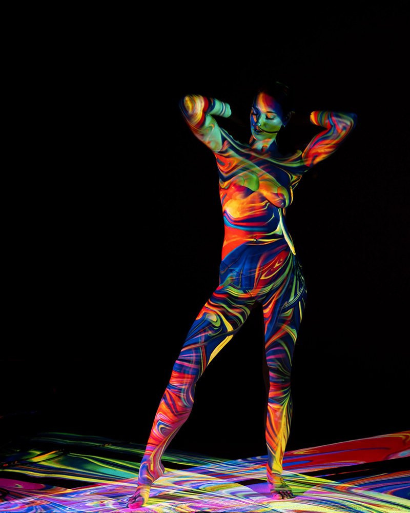 A person standing with arms bent behind the head, covered in swirling, vibrant multicolored patterns projected onto their body and the floor against a black background