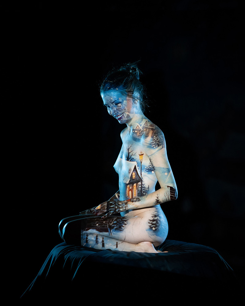 Nude woman sitting cross-legged on a black surface, with a winter village scene projected onto her body, including snow-covered houses, trees, and a street lamp, against a black background.