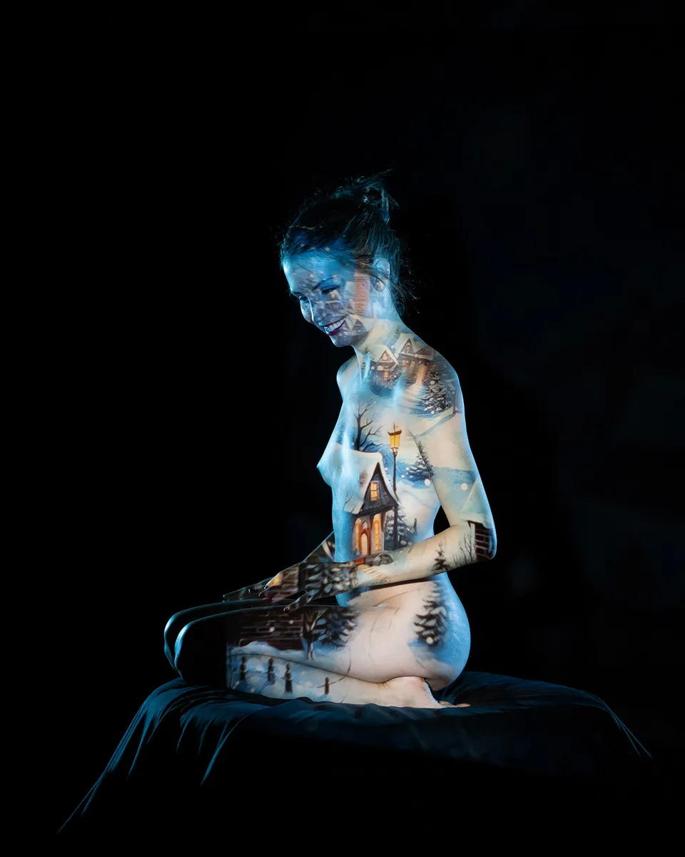 Nude woman sitting cross-legged on a black surface, with a winter village scene projected onto her body, including snow-covered houses, trees, and a street lamp, against a black background.