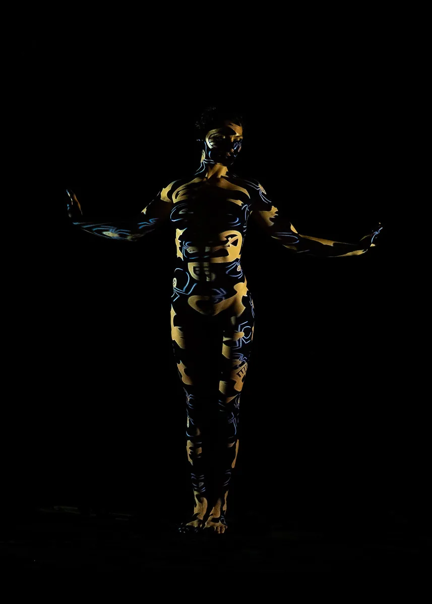 Person standing with arms extended, covered in glowing yellow and blue abstract patterns on their body against a black background