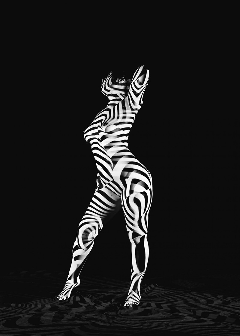 Nude person standing on one foot with arms raised behind head, covered in black and white zebra-like stripes projected against a black background