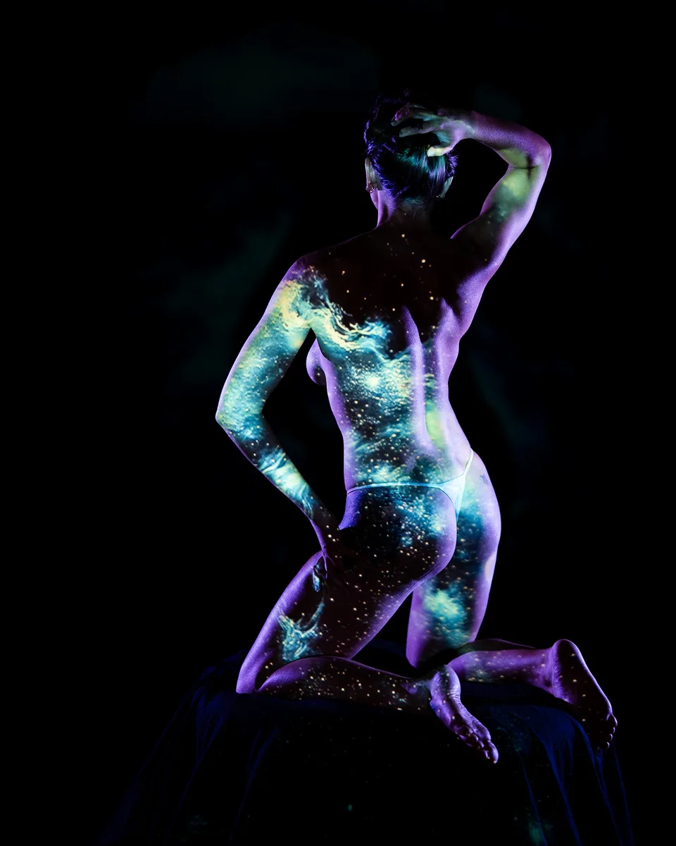 A figure kneeling on a dark surface with one hand resting on their head, their body illuminated by a projected pattern of swirling blue and purple cosmic light and stars, creating an intimate and contemplative mood.
