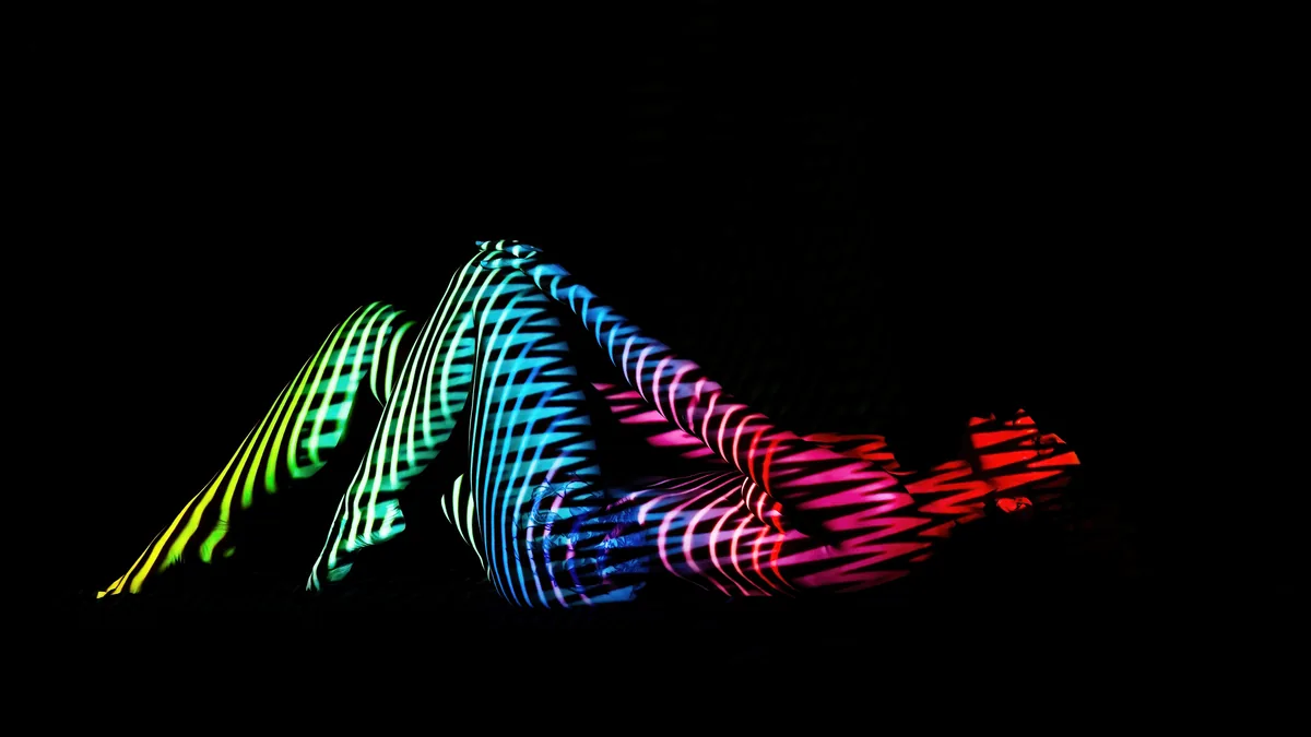 A person reclines against a black background with blue, green, and red striped light patterns projected onto their body, creating a geometric interplay of color and shadow.