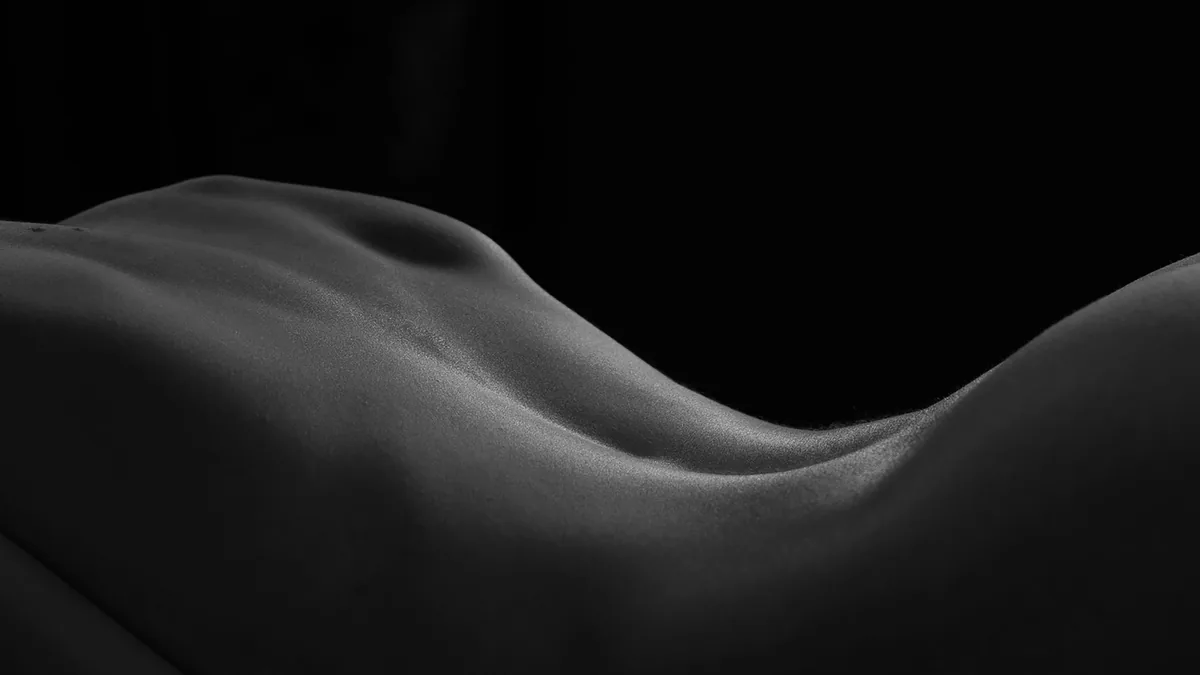 Close-up of a smooth human back with gentle highlights and shadows emphasizing the spinal curve and muscle contours against a dark background