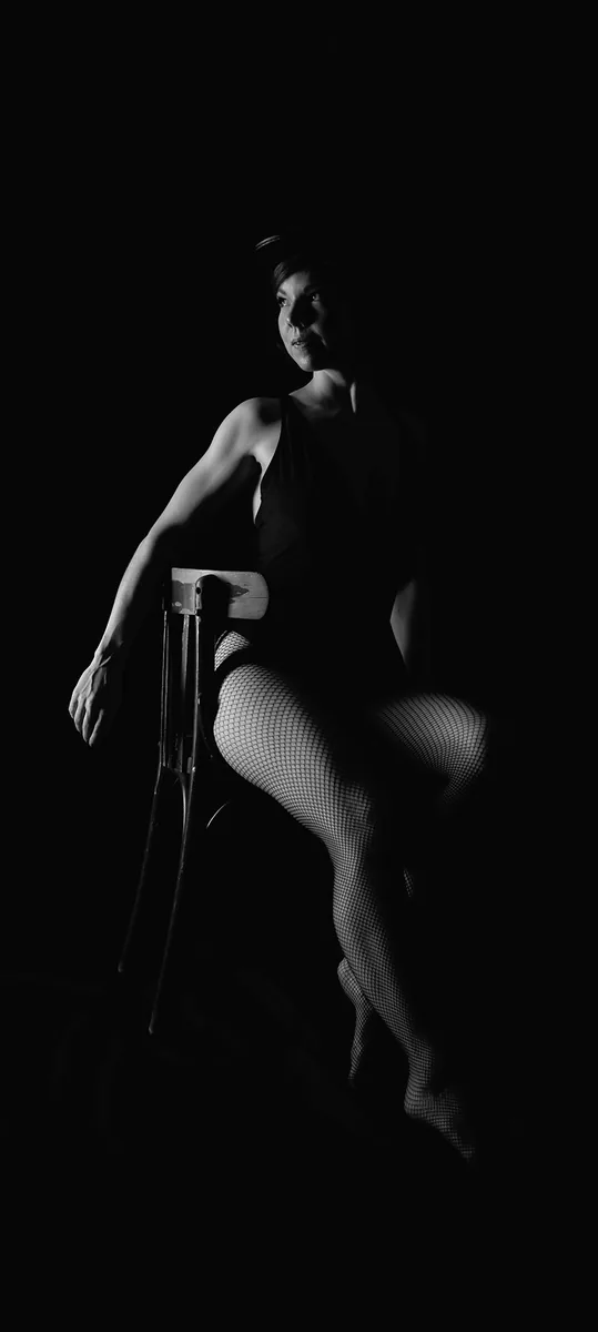 Person sitting on wooden chair wearing a sleeveless outfit, fishnet tights, and a hat, with dramatic side lighting highlighting their face, arm, and legs against a black background.
