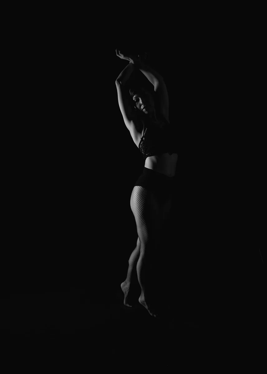 Woman posing with arms raised and legs crossed, wearing fishnet stockings and dark lingerie, illuminated against a black background