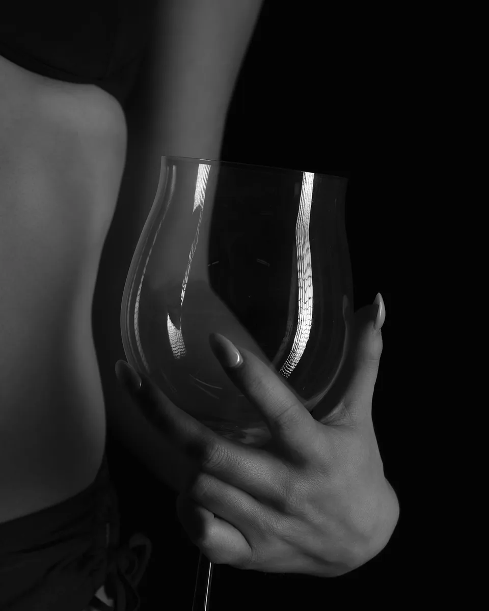 Close-up of a hand with long fingernails holding an oversized empty wine glass against a dark background; part of a bare torso is visible on the left side