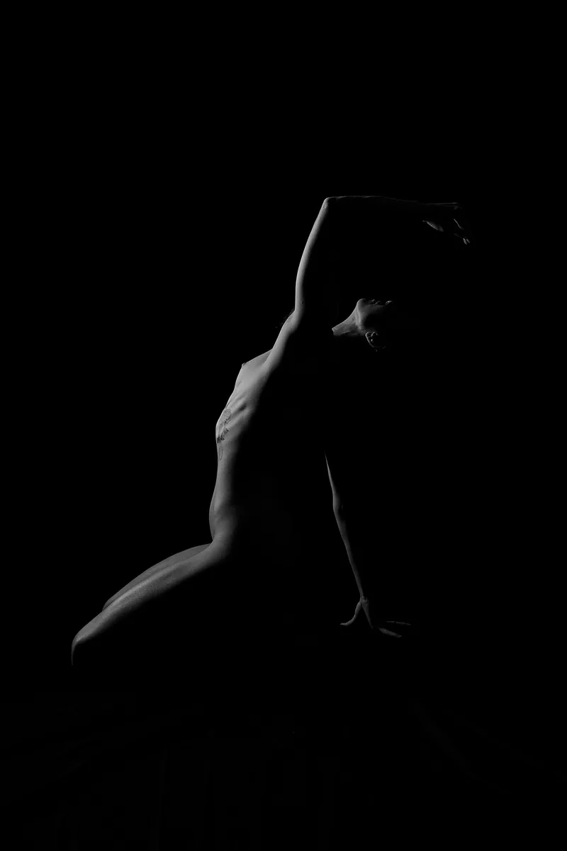 Side view of a person kneeling and arching their back, with one arm extended over their head, captured as a silhouette against a black background.